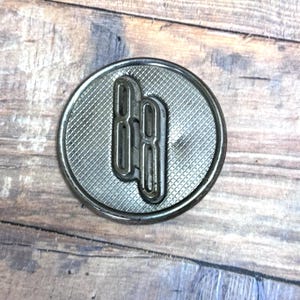 May include: A circular, silver-toned metal emblem with a textured background and a raised, stylized "88" design. The emblem is set against a weathered, wooden surface, showcasing its metallic sheen.