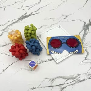 May include: A game set including a yellow, green, red, and blue textured plastic cluster, a white die with the text I choose, a white notepad, and a blue decoder mask with red lenses and the text Cranium Cadoo Decoder Mask printed on the front.