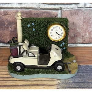 Puede incluir: Un reloj decorativo con temática de golf que presenta un carrito de golf en miniatura, una bolsa de golf y un reloj con borde dorado. El reloj tiene una esfera blanca con números y manecillas negras. La base es verde.