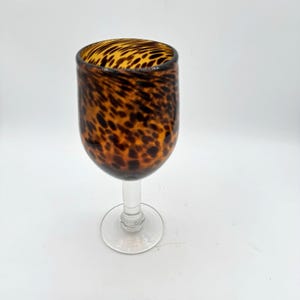 Puede incluir: Una copa de cristal con un cuenco redondeado que presenta un patrón de manchas de animal en tonos marrones y negros. El tallo y la base circular están hechos de vidrio transparente, lo que contrasta con la parte superior estampada del recipiente.