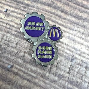 May include: A metal pin in the shape of three interlocking gears. The top gear has the text "GO GO GADGET" in gold on a purple background. The middle gear has the McDonald's logo. The bottom gear has the text "CODE NAME GAME".