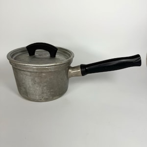 May include: A small, vintage aluminum saucepan with a lid and a long, black handle. The pot has a rounded body and a lid with a black handle. The handle is curved and attached to the side of the pot.