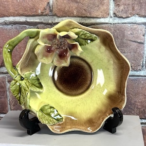 May include: A decorative ceramic dish with a floral design. The dish features a raised flower with yellow petals and a brown center. The handle and leaves are green. The dish is supported by a black stand.