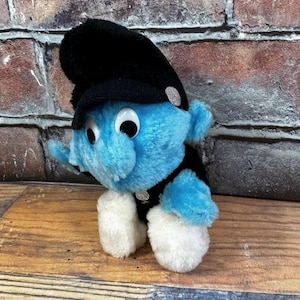 May include: A plush blue Smurf toy wearing a black hat and jacket with silver accents. The toy has large, round eyes and white fluffy feet. The background features a brick wall and wooden surface.