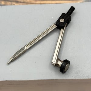Pode incluir: Um compasso prateado e preto, ferramenta para desenhar círculos. Possui duas pernas ajustáveis, uma ponta afiada e uma roda de ajuste preta. Este instrumento de precisão é usado em geometria e desenho técnico, com medidas em centímetros.
