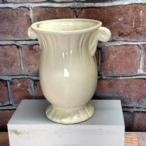 Puede incluir: Un jarrón de cerámica de color crema con dos asas. El jarrón tiene un diseño acanalado y una base redonda.