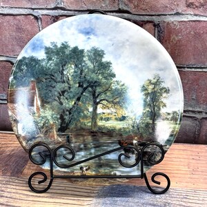 May include: Decorative plate featuring a landscape painting with a river, trees, and a small house. The plate is displayed on a black metal stand. The artwork has a vintage aesthetic with muted colors.