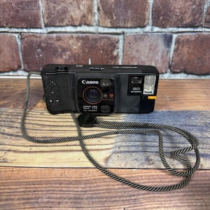 May include: A black Canon Snappy 20 camera with a black and gray strap. The camera has a Canon lens and auto focus features. The camera is on a wooden surface with a brick background.