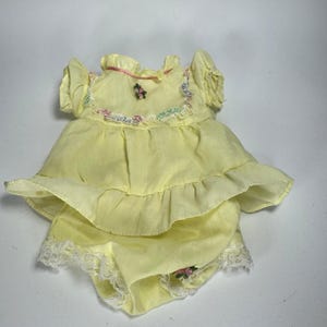May include: A yellow doll dress and matching bloomers set. The dress features a ruffled skirt, short sleeves, and floral embroidery. The bloomers have a lace trim. The set is made of a lightweight fabric.
