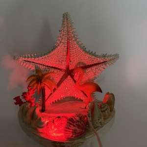Pode incluir: Lâmpada decorativa com tema de praia, com uma grande estrela do mar branca brilhante e iluminação interna vermelha. A lâmpada inclui um flamingo, uma palmeira e conchas, criando um ambiente tropical. A base tem formato de concha.