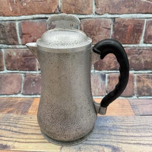 May include: A silver-toned metal coffee pot with a textured, speckled finish. The pot features a matching lid with a decorative handle and a curved, black heat-resistant handle attached to the side. The body has a wide base that tapers upward.