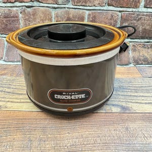 May include: A Rival Crock-Ette slow cooker with a dark brown cylindrical body, a glossy brown rim, and a black lid. The front features a label with the text RIVAL CROCK-ETTE SLOW COOKER and a small orange indicator light near the base.