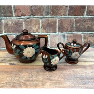 May include: A brown ceramic tea set consisting of a teapot with a lid, a creamer, and a sugar bowl with a lid. Each piece features a hand-painted design of large cream-colored flowers with teal green leaves against a dark brown glazed finish.