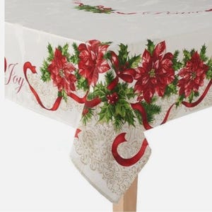 Puede incluir: Un mantel navideño blanco con un diseño festivo. El borde presenta flores de pascua rojas, acebo verde y cintas rojas. La palabra "Joy" está escrita en cursiva en el mantel.