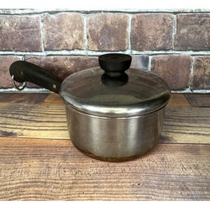 May include: A stainless steel saucepan with a black handle and lid. The handle has a metal ring. The pot is on a wooden surface. The background is a brick wall.