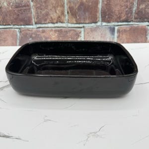 May include: A rectangular ceramic dish with a glossy black finish and rounded corners. The dish features a deep, open basin with smooth, reflective surfaces and a solid, uniform dark color throughout the entire exterior and interior structure.
