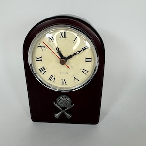Puede incluir: Un reloj de cuarzo de madera marrón oscuro con esfera crema y números romanos. El reloj tiene un bisel plateado y un segundero rojo. Una pelota de golf y palos de golf cruzados están representados.
