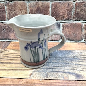 May include: A light gray ceramic pitcher with a wide spout and a curved handle. The exterior features a hand-painted design of blue iris flowers with green stems above a brown horizontal band. A small rectangular label reads Reynolds Studio Lansing, Michigan.