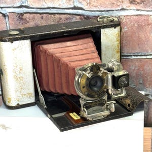 May include: A vintage black and silver folding camera with a red bellows and a lens with a silver cap. The camera has a leather strap and a small viewfinder on the side.