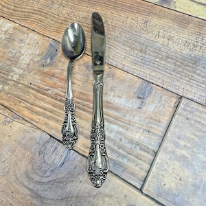 以下が含まれることがあります： 装飾的なハンドルが付いた銀のスプーンと銀のバターナイフ。食器は木製の表面の上にあります。