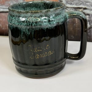 May include: A dark green and black ceramic mug with a handle. The top rim has a green glaze that drips down the sides. The words "Quebec Canada" are written in gold script on the front of the mug.