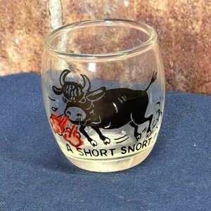 以下が含まれることがあります： 黒い雄牛のイラストと「A SHORT SNORT」の文字が入った透明なショットグラス。雄牛は口に赤い物体を持っています。グラスは青い表面に置かれています。