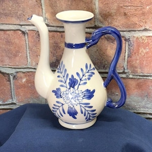 Può includere: Una teiera in ceramica bianca con un motivo floreale blu. La teiera ha un beccuccio lungo e un manico che si curva all'indietro.