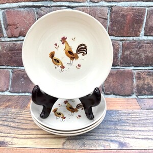 以下が含まれることがあります： 中央に茶色と金色の雄鶏と雌鶏のイラストが描かれた、クリーム色の陶器製ボウルセット。ボウルは積み重ねられ、濃い茶色のプラスチック製スタンドで支えられており、各ピースには農場の鳥のモチーフが繰り返しデザインされています。