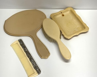 Può includere: Un set da toeletta vintage con uno specchio da mano, una spazzola, un pettine e un portasapone. Il set è realizzato in plastica color avorio.