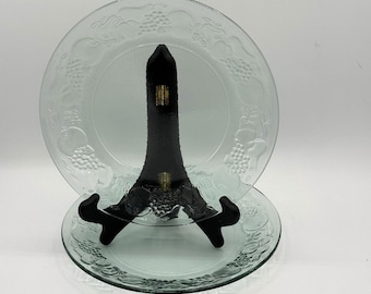May include: Two clear glass plates with a fruit design, displayed on a black plate stand. The plates have a raised pattern of grapes and other fruits around the rim. The plates are round and appear to be about 10 inches in diameter.