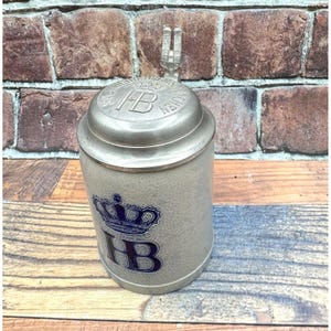 May include: A beige ceramic beer stein featuring a blue crown and the letters HB on the front. The stein has a metallic lid with an embossed crown, the letters HB, and a small metal figurine of a building on top of the lid hinge.