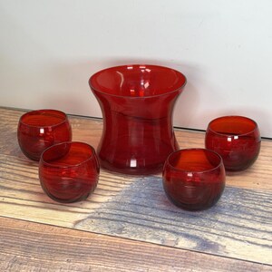 Puede incluir: Conjunto de cinco vasos de vidrio rojo, que incluyen un vaso más grande en forma de cuenco y cuatro vasos redondos más pequeños. Los vasos son todos de vidrio transparente con un tinte rojo.