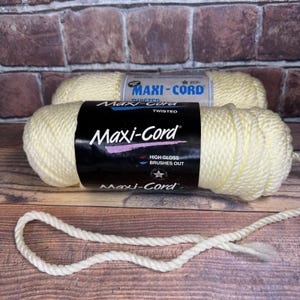 May include: Two spools of off-white Maxi-Cord twisted yarn are displayed, with one in the foreground and another behind it. The front spool's label is black with white and pink text. A length of the yarn is unraveled, showing its texture.
