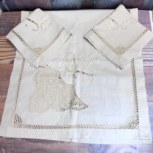 Puede incluir: Un juego de mesa de lino color crema con un mantel rectangular y dos servilletas cuadradas a juego. El juego presenta intrincados calados y diseños florales bordados. Los bordes del mantel y las servilletas están decorados con un delicado encaje.