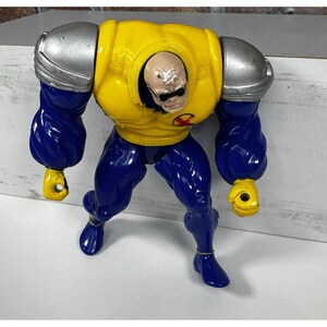 May include: A plastic action figure of a character with a bald head, wearing sunglasses, and a yellow and blue uniform. The figure has large silver shoulder pads and yellow hands. The character's legs are blue, and the figure is posed in a dynamic stance.