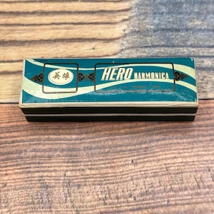 May include: A vintage harmonica box with the text "HERO HARMONICA" in white lettering. The box is teal with a black base and a white wave design. The box also has Chinese characters.