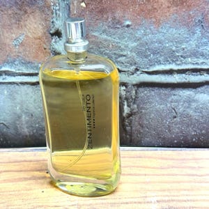 May include: A clear glass perfume bottle with a silver cap, filled with a yellow liquid. The bottle has a rectangular shape with rounded edges. The word "Zentimento" is printed vertically on the front, along with "fragrance collection".