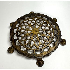 May include: Ornate, round, metal trivet with a gold and bronze finish. The trivet features a detailed, openwork design with a central starburst pattern and decorative feet. This vintage-style piece is designed to protect surfaces from heat.