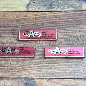 Puede incluir: Tres cajas vintage rojas y blancas de Mir A Kut Co. de cuchillas Super Stainless Steel Shaper. Las cajas tienen forma rectangular y están dispuestas sobre una superficie de madera.