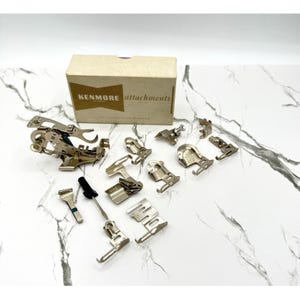 Puede incluir: Una colección de accesorios para máquinas de coser Kenmore, que incluye varios prensatelas y un destornillador, dispuestos sobre una superficie de mármol blanco. Una caja de cartón con el logotipo de Kenmore y la palabra "attachments" está en el fondo.