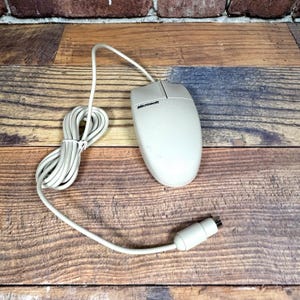 May include: Off-white Microsoft computer mouse with a cord and connector. The mouse has a classic design with a scroll wheel and the Microsoft logo. The cord is coiled and the mouse is on a wooden surface.