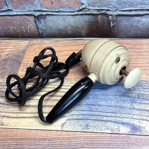 May include: An antique, cream-coloured electric hair dryer with a black handle and a white, circular end piece. The dryer has a coiled black cord and a black plug. The background is a mix of wood and brick.