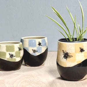 May include: Three ceramic planters with a rounded shape. Each features a black base and a checkered upper section in sage green, light blue, or pale yellow. Black bee illustrations are printed on the checkered areas. One planter contains a green plant.