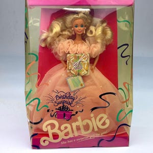 Puede incluir: Una muñeca Barbie rubia con un vestido color melocotón con escote de volantes, empaquetada en una caja con la etiqueta Birthday Surprise. La caja presenta gráficos de cintas de colores, un accesorio de regalo y el texto She has a surprise gift for you!