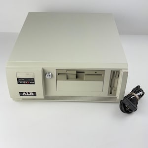 Puede incluir: Una torre de computadora de escritorio beige con el logotipo de la marca ALR, una etiqueta VEISA ALR, una cerradura circular, una disquetera horizontal y una disquetera vertical. Un cable de alimentación negro está atado con una banda elástica.