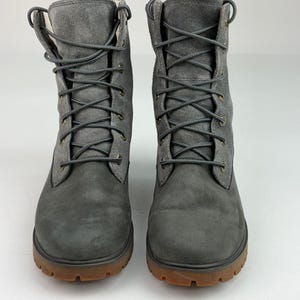Puede incluir: Un par de botas con cordones a media pantorrilla en un acabado gris carbón mate. Las botas presentan una puntera de cuero liso, una caña con textura de ante, cordones grises a juego, ojales metálicos y una suela de goma gruesa y resistente.