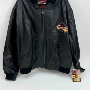 May include: A black leather bomber jacket featuring a ribbed collar, cuffs, and hem. The jacket has a front zipper closure, two side pockets, and a Corvette logo patch on the left chest. A Reed Leather Sportswear brand tag is attached to the sleeve.