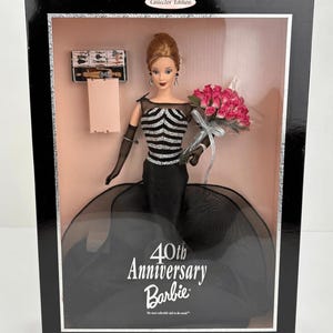 Puede incluir: Una muñeca Barbie 40th Anniversary con un vestido de corpiño a rayas negras y plateadas, guantes negros transparentes y un ramo de rosas rosas. La muñeca se presenta en una caja negra con el texto 40th Anniversary Barbie y Collector Edition.