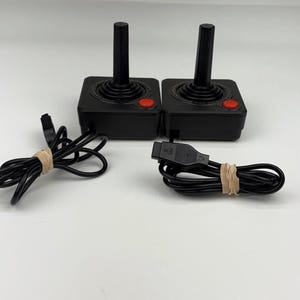 Puede incluir: Dos mandos de videojuegos negros con base cuadrada, un joystick vertical y un único botón rojo en la esquina superior derecha. Cada mando cuenta con un cable negro con un conector rectangular, enrollado y sujeto con una goma elástica beige.
