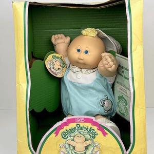 May include: A 1985 Cabbage Patch Kids Preemie doll with blonde hair and blue eyes, wearing a light blue romper with an elephant graphic and a white collared shirt. The doll is inside its original yellow and green cardboard packaging box.